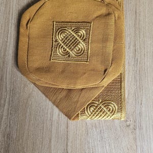 May include: Mustard-colored fabric item with an embroidered gold design. The item has a rounded shape with a square embroidered detail in the center. The item is placed on a wooden surface.
