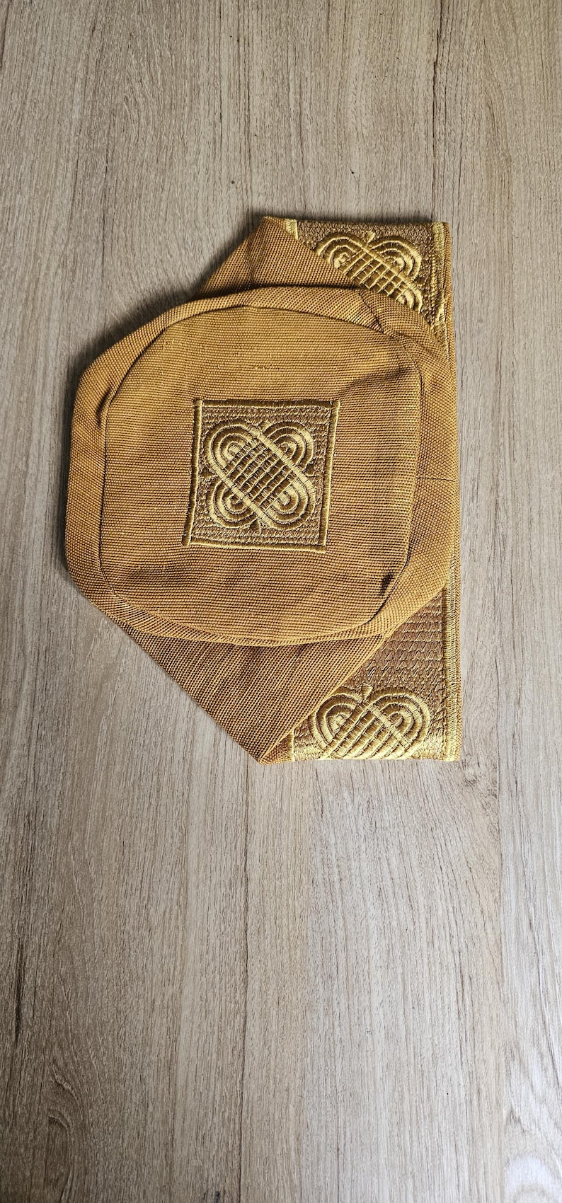May include: Mustard-colored fabric item with an embroidered gold design. The item has a rounded shape with a square embroidered detail in the center. The item is placed on a wooden surface.