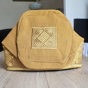 May include: A gold-colored, structured hat with an intricate embroidered design on the front and sides. The hat features a square, embroidered emblem in the center and decorative patterns along the edges. The fabric has a subtle texture.