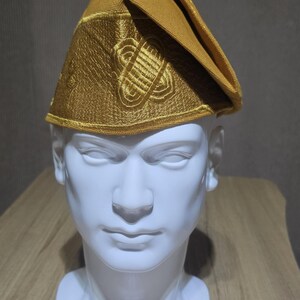 May include: A gold-colored hat with intricate embroidered designs, displayed on a white mannequin head. The hat has a structured shape with a folded brim and detailed patterns. The background is a neutral gray.