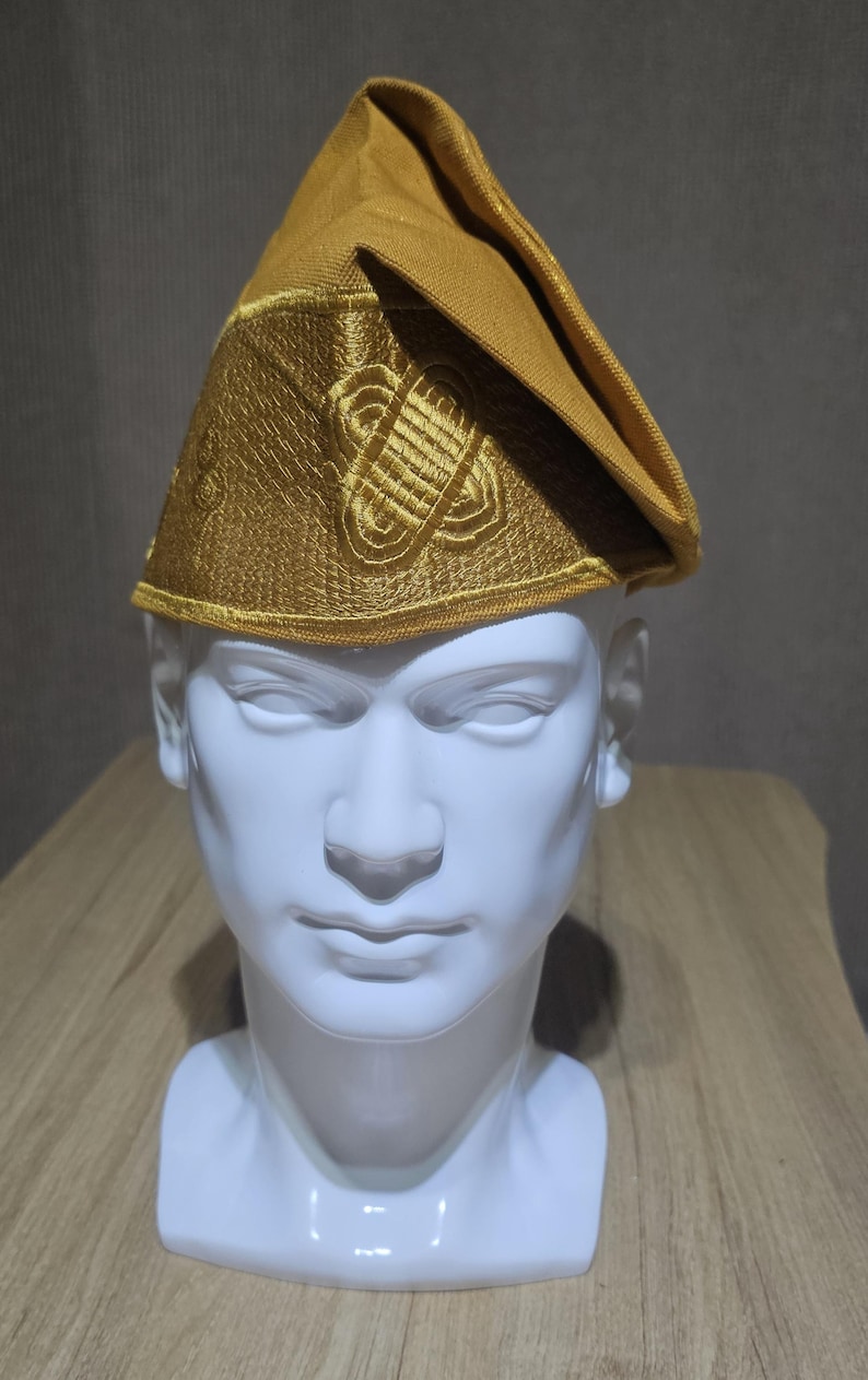 May include: A gold-colored hat with intricate embroidered designs, displayed on a white mannequin head. The hat has a structured shape with a folded brim and detailed patterns. The background is a neutral gray.