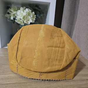 Può includere: Un cappello intrecciato dorato con un bordo cucito. Il cappello ha una parte superiore arrotondata e un bordo piatto.