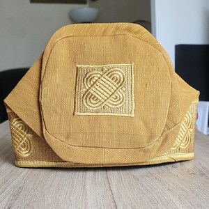 May include: A gold-colored, structured hat with an intricate embroidered design on the front and sides. The hat has a square embroidered patch in the center and decorative patterns along the edges. The fabric appears to have a subtle texture.
