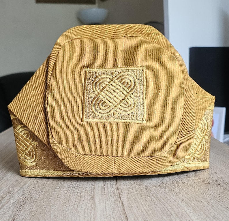 May include: A gold-colored, structured hat with an intricate embroidered design on the front and sides. The hat has a square embroidered patch in the center and decorative patterns along the edges. The fabric appears to have a subtle texture.