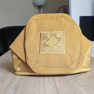 May include: A gold-colored hat with an embroidered square design on the front. The hat has a structured shape with decorative embroidery along the edges. The hat is made of a textured fabric and sits on a wooden surface.