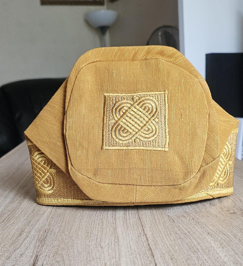 May include: A gold-colored hat with an embroidered square design on the front. The hat has a structured shape with decorative embroidery along the edges. The hat is made of a textured fabric and sits on a wooden surface.