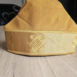 May include: A gold-colored, traditional hat with a rectangular crown and a decorative band. The band features an embroidered pattern with a repeating knot design. The hat is likely used for cultural or ceremonial purposes.