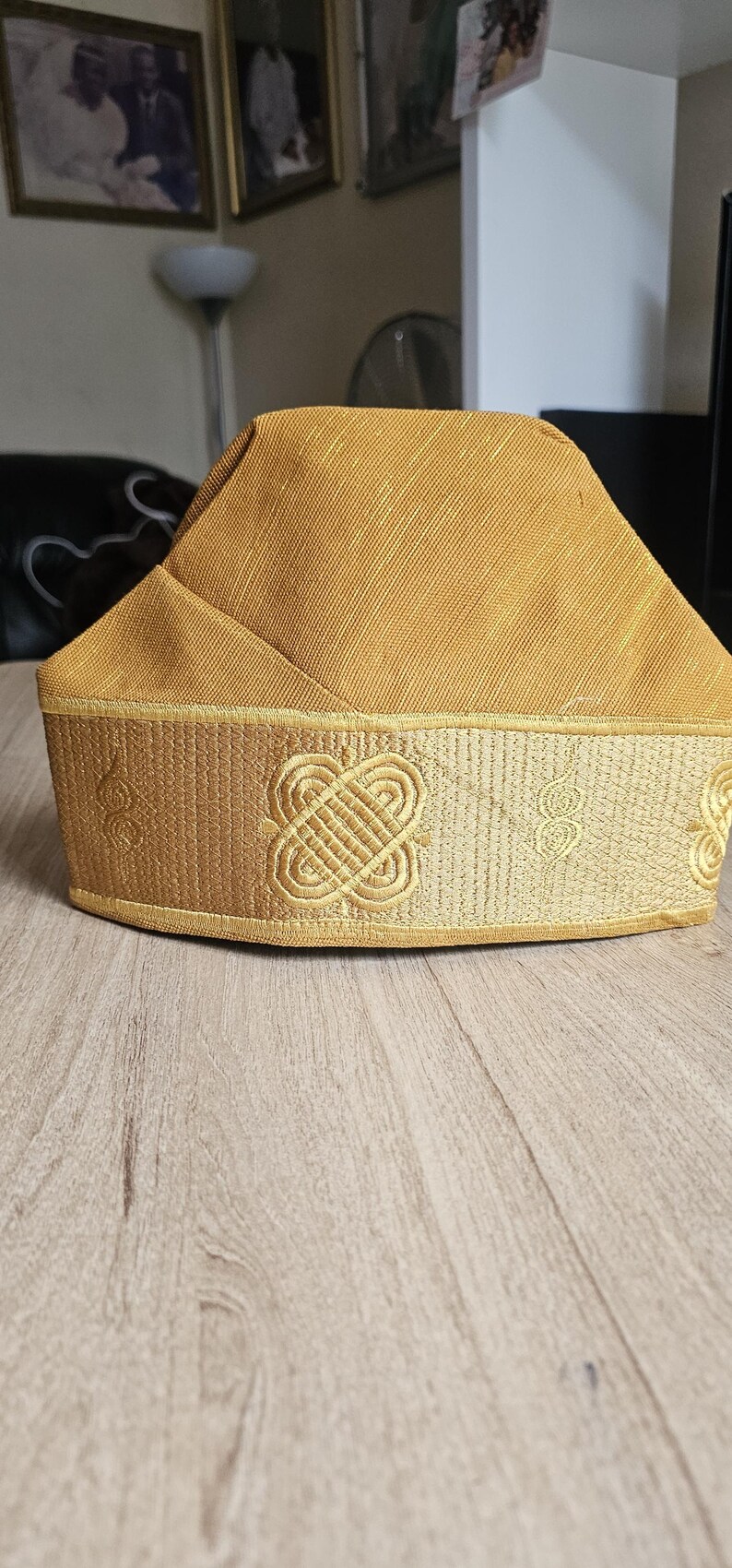 May include: A gold-colored, traditional hat with a rectangular crown and a decorative band. The band features an embroidered pattern with a repeating knot design. The hat is likely used for cultural or ceremonial purposes.