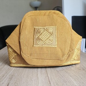 May include: A gold-colored, structured hat with intricate embroidered designs. The hat features a square emblem with a detailed pattern and additional embroidery along the edges. The fabric has a subtle texture, and the hat sits on a wooden surface.