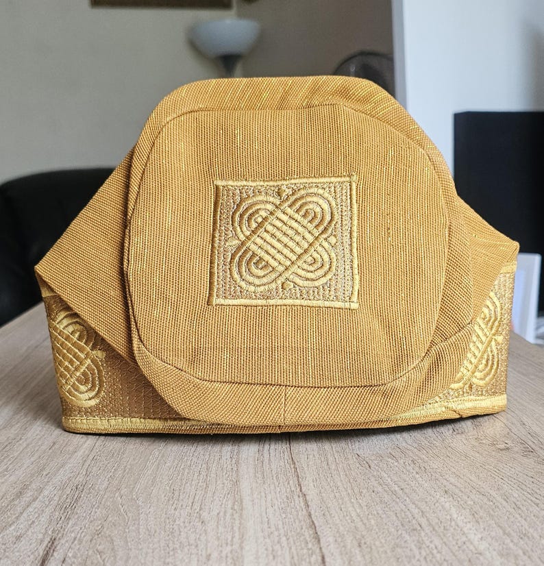 May include: A gold-colored, structured hat with intricate embroidered designs. The hat features a square emblem with a detailed pattern and additional embroidery along the edges. The fabric has a subtle texture, and the hat sits on a wooden surface.