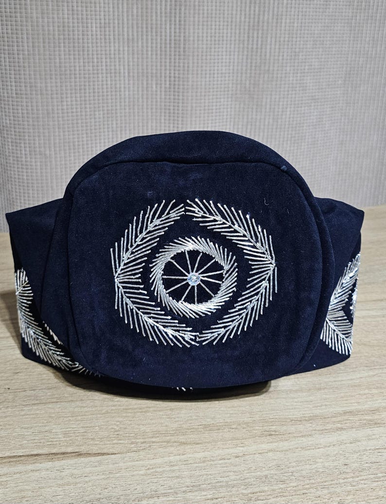 May include: A dark blue hat with a circular design embroidered in silver thread. The design features a central circle with radiating lines.