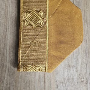 May include: A gold-colored, padded fabric item with an embroidered design. The embroidered section features a repeating pattern of interlocking shapes and lines. The item is set against a light wood grain background.