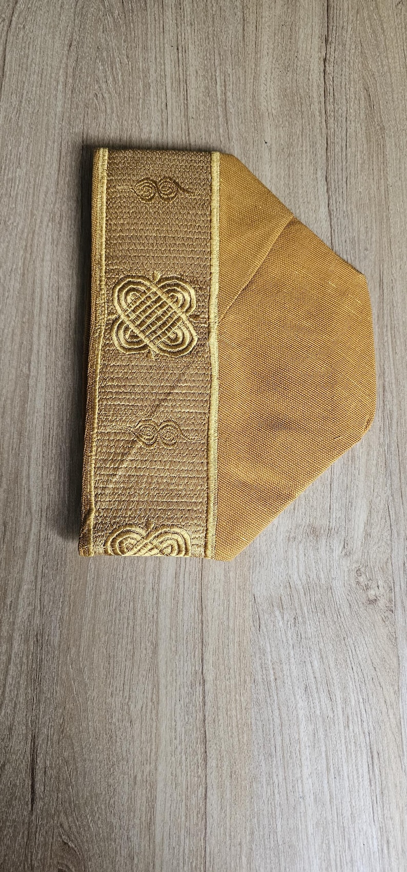 May include: A gold-colored, padded fabric item with an embroidered design. The embroidered section features a repeating pattern of interlocking shapes and lines. The item is set against a light wood grain background.