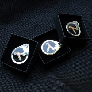 Puede incluir: Tres pines de metal en forma de lágrima con un centro circular negro, un símbolo lambda griego de color cobre y el número 3 en blanco. Cada pin se presenta dentro de una caja de regalo cuadrada individual de color negro con acabado mate.