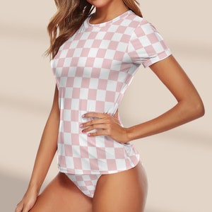May include: Pink and white checkered swimsuit top with a scoop neck and short sleeves. The top is paired with matching checkered bikini bottoms. The brand name "Lucky Suzy Beachwear" is printed on a pink circle with a blue circle inside.