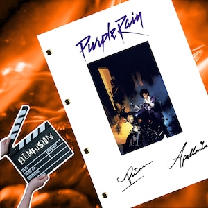 May include: A white sheet of paper with the title "Purple Rain" written in purple letters. A black and white photo of Prince on a motorcycle is in the center of the page. The photo is signed by Prince and Apollonia.