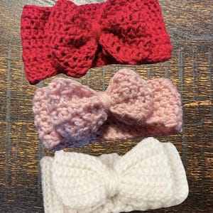 May include: Three handmade crochet headbands in red, light pink, and white. Each headband features a large bow in the center. The headbands are made of soft yarn and are suitable for babies and toddlers.