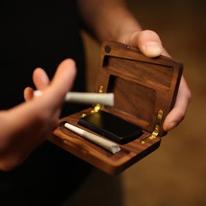 May include: A wooden box with a hinged lid, containing two rolled cigarettes and a small black rectangle. The box is made of dark wood with gold hinges.