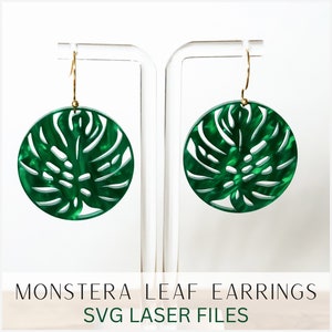 Könnte beinhalten: Ein Paar grüne Monstera-Blattohrringe mit goldenen Haken. Die Ohrringe sind aus lasergeschnittenem Acryl und werden auf einem weißen Ständer präsentiert. MONSTERA LEAF EARRINGS SVG LASER FILES