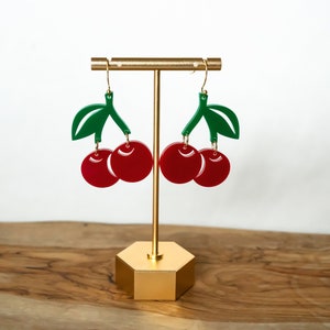 Op de afbeelding: Een paar rode kersenoorringen met groene bladeren die aan een gouden standaard hangen. De oorbellen zijn gemaakt van acryl en hebben een glanzende afwerking.