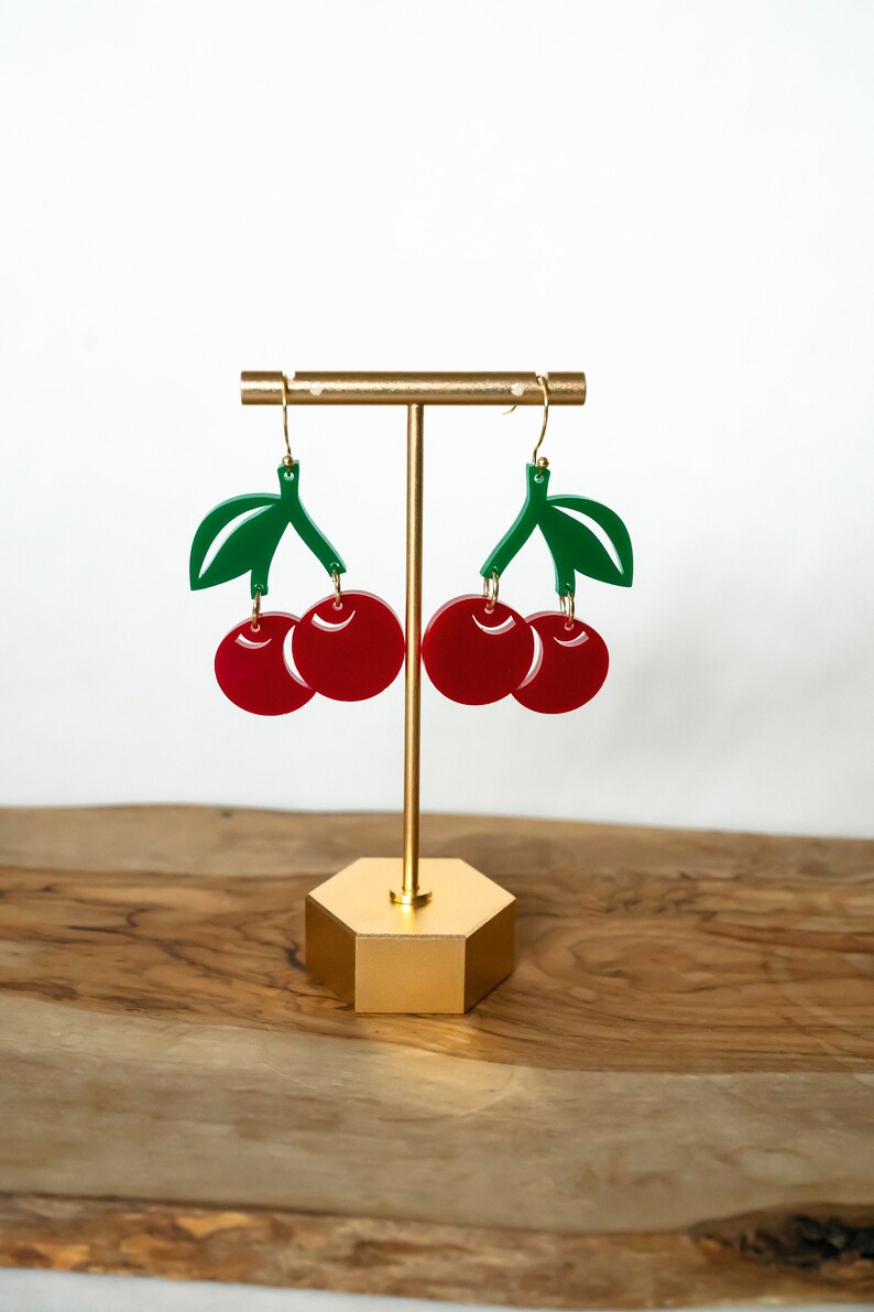 Op de afbeelding: Een paar rode kersenoorringen met groene bladeren die aan een gouden standaard hangen. De oorbellen zijn gemaakt van acryl en hebben een glanzende afwerking.