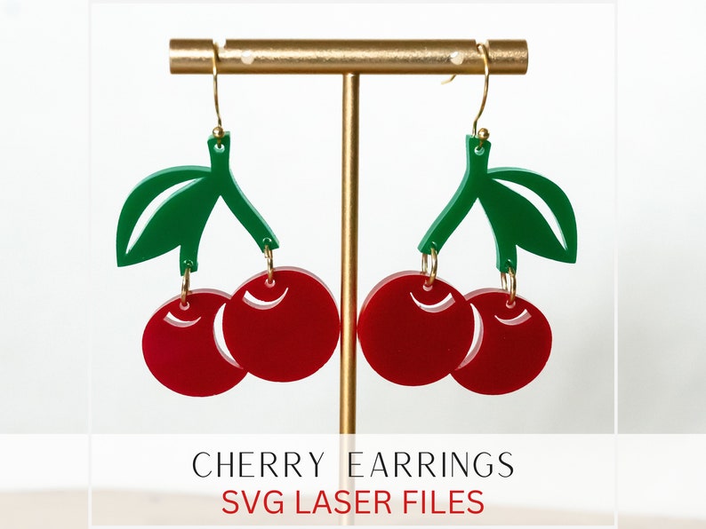 Op de afbeelding: Een paar rode en groene kersenoorringen op een gouden standaard. De oorbellen zijn gemaakt van acryl en hebben een glanzende afwerking.  "CHERRY EARRINGS SVG LASER FILES" staat onder de oorbellen.