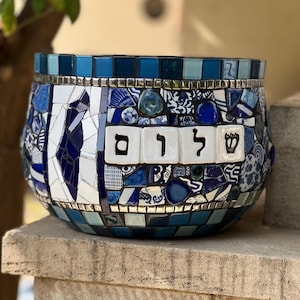 Puede incluir: Una maceta de mosaico con una combinación de colores azul y blanco. La maceta presenta una variedad de azulejos azules y piezas de cerámica, con la palabra hebrea "Shalom" en la parte delantera. La maceta está sobre una repisa de piedra.