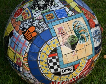 May include: A mosaic sphere with a variety of colorful tiles. The sphere features images of a rooster, a bee, and the words "HOME", "IS HERE", and "OMG". The sphere is resting on green grass.