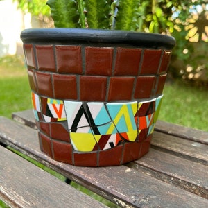 Può includere: Un vaso in ceramica color terracotta con un bordo nero, decorato con un mosaico di forme geometriche colorate. Il vaso è appoggiato su una superficie di legno.