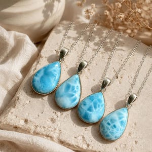 May include: Four silver necklaces with teardrop-shaped pendants. Each pendant features a light blue gemstone with white marbling, set in a silver bezel. The necklaces are displayed on a textured, light-colored surface.