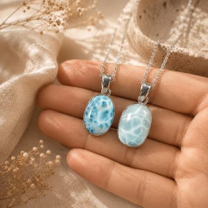 May include: Two oval-shaped, light blue larimar stone pendants on silver chains. The pendants have a marbled appearance, with varying shades of blue and white. The necklaces are displayed on a hand, with a neutral-toned background.