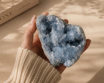 May include: A light blue celestite crystal geode held in a hand. The crystal has a rough, textured exterior and a cluster of sparkling blue crystals inside. The geode is a natural, organic shape, and the color is a soft, pale blue.