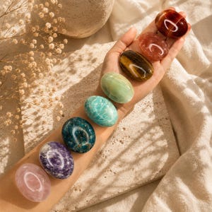 May include: Seven polished gemstones in shades of pink, purple, teal, green, brown, and red. The oval-shaped stones are displayed on a light surface. The gemstones are held in a hand.