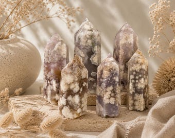 May include: Six polished crystal towers in shades of lavender, cream, and brown are arranged on a textured stone surface. The crystals vary in size and shape, with pointed tops and visible internal patterns. Dried floral accents and a neutral background complete the display.