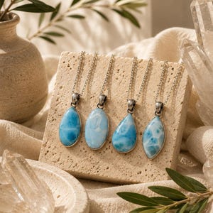 May include: Four silver necklaces featuring light blue larimar pendants in a teardrop shape. The pendants have a marbled effect and are displayed on a textured, beige stone. The necklaces are on a beige surface with decorative elements.