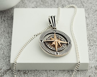 May include: Silver-toned compass pendant necklace with a gold-colored compass rose. The pendant features directional markings and is suspended from a delicate silver chain. The necklace is displayed on a white surface against a neutral background.