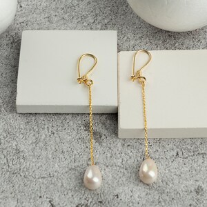 May include: A pair of gold-colored drop earrings. Each earring features a gold-colored hook, a delicate chain, and a teardrop-shaped, off-white pearl. The earrings are displayed on a white surface against a neutral background.