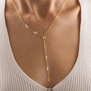 May include: A gold-colored necklace with the name "Jennifer" spelled out in individual letters. The necklace has a delicate chain and a Y-shaped design, with the letters hanging vertically. The necklace is worn on a person.