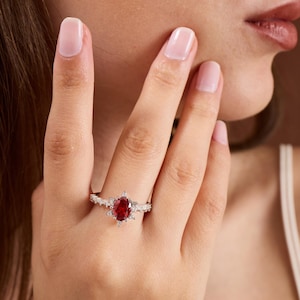 May include: A silver ring with an oval, deep red gemstone as the centerpiece. The ring is adorned with smaller, clear gemstones around the main stone and along the band. The ring is on a person's finger.