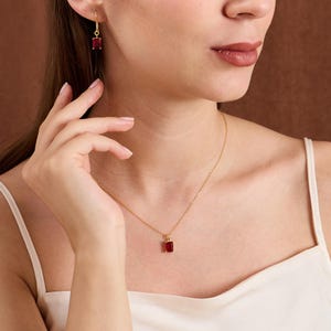 May include: A gold necklace and earrings set featuring rectangular, deep red gemstones. The necklace pendant and earring drops are suspended from delicate gold chains and hooks. The jewelry is worn against a neutral background.