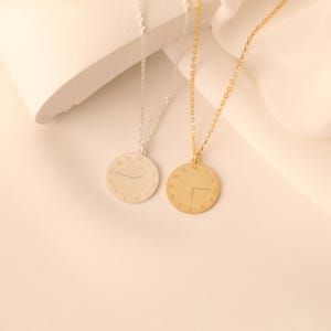 Op de afbeelding: Twee kettingen met ronde hangers, één zilver en één goud. Elke hanger heeft een wijzerplaatontwerp met de cijfers 1 tot en met 12. De zilveren ketting heeft een eenvoudige lijn voor de wijzers, terwijl de gouden ketting de wijzers op 2:45 uur toont.