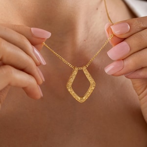 Puede incluir: Un collar dorado con un colgante en forma de diamante. El colgante presenta un centro abierto con un diseño ornamental y ondulado. El collar lo sostiene una persona con uñas cuidadas.