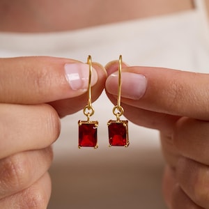 May include: A pair of gold-colored drop earrings, each featuring a rectangular, deep red gemstone. The earrings are held up against a neutral background, showcasing their vibrant color and design. The gemstones are approximately 0.5 inches in length.