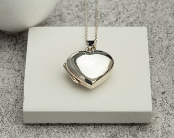 Puede incluir: Un colgante de locket en forma de corazón de plata en una cadena de plata. El locket está cerrado y tiene una superficie lisa y reflectante.