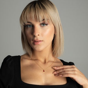 May include: A woman with blonde hair wearing a black top and a gold necklace with a red gemstone pendant.