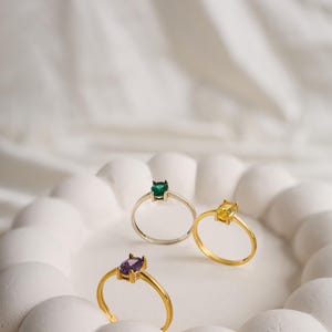 Peut inclure: Trois bagues en or avec des pierres précieuses de différentes couleurs. Les bagues sont sur une surface blanche avec un motif texturé.