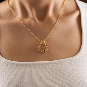 Puede incluir: Un collar de tono dorado con una cadena fina y un colgante en forma de gota abierta y entrelazada. El colgante está decorado con una pequeña piedra preciosa de color violeta intenso situada en la base del bucle, creando una pieza sencilla.