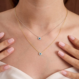 May include: Two layered necklaces, one gold-toned and one silver-toned, each with a small, round pendant with a blue eye design. The necklaces are displayed against a neutral background, creating a simple yet elegant aesthetic. The pendants are small and delicate.