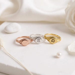 Könnte beinhalten: Drei Siegelringe in einer Reihe angeordnet, bestehend aus einem roségoldenen Ring mit leerer runder Fläche, einem silbernen Ring mit der Gravur des Buchstabens A und einem goldenen Ring mit der Gravur des Buchstabens D. Jeder Ring hat ein strukturiertes Band.