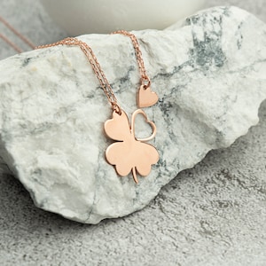 Könnte beinhalten: Eine Halskette aus Roségold mit einem vierblättrigen Kleeblatt-Anhänger und einem kleineren Herz-Anhänger. Die Kette liegt auf einem weißen Stein.